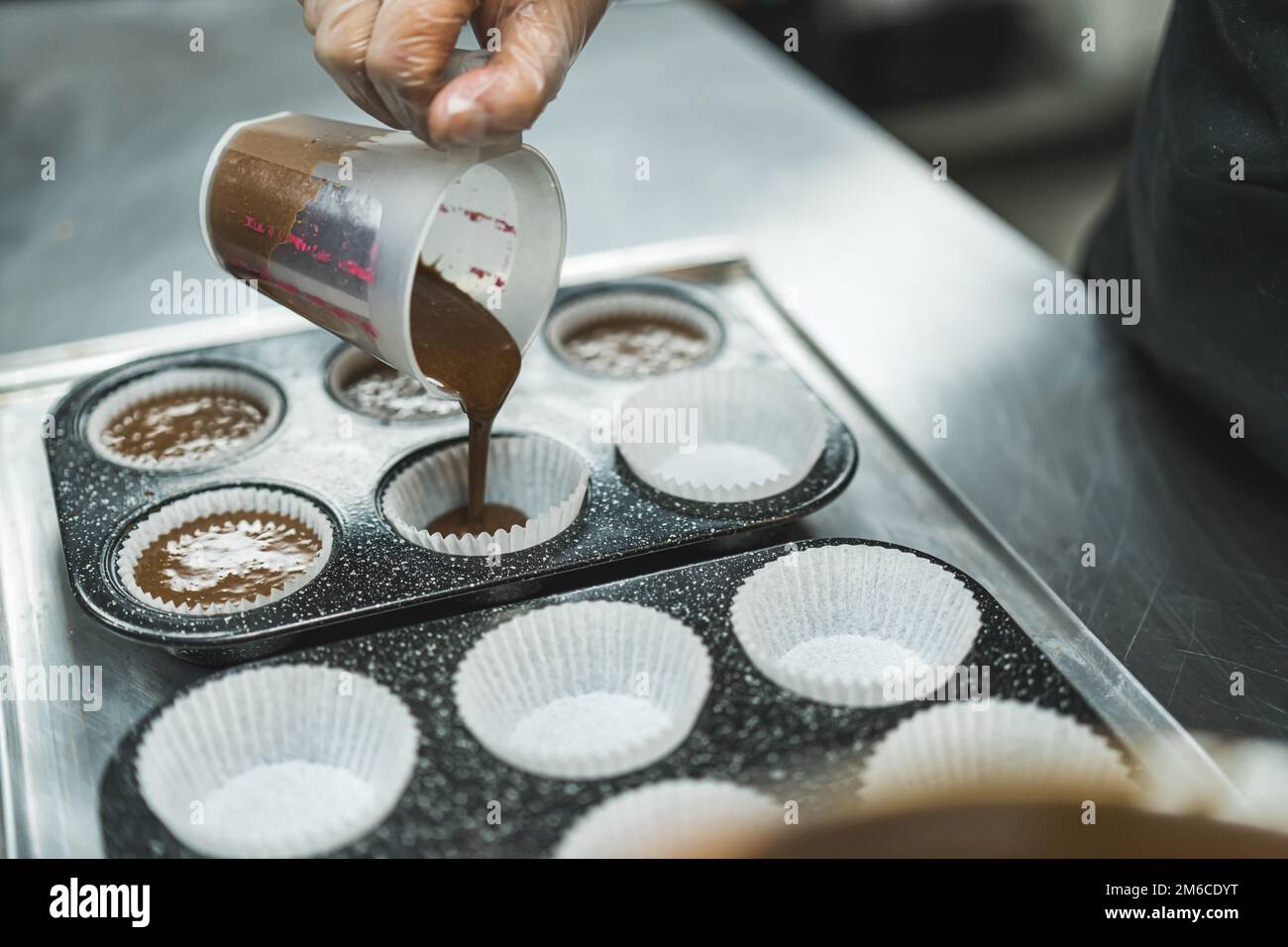 Vista laterale dell'impasto di cioccolato versato nelle fodere dei cupcake in due padelle per cupcake da un panettiere che indossa un guanto trasparente. Foto di alta qualità Foto Stock