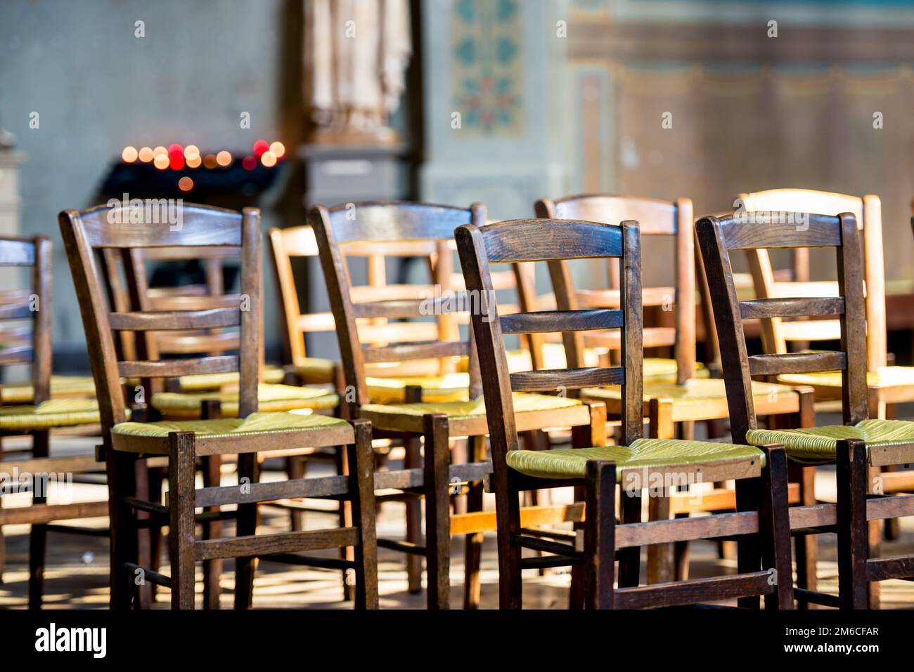 Una vista ravvicinata delle sedie in legno con posti a sedere in vimini verdi allineati davanti ad una rastrelliera che tiene accese candele votive. Foto Stock
