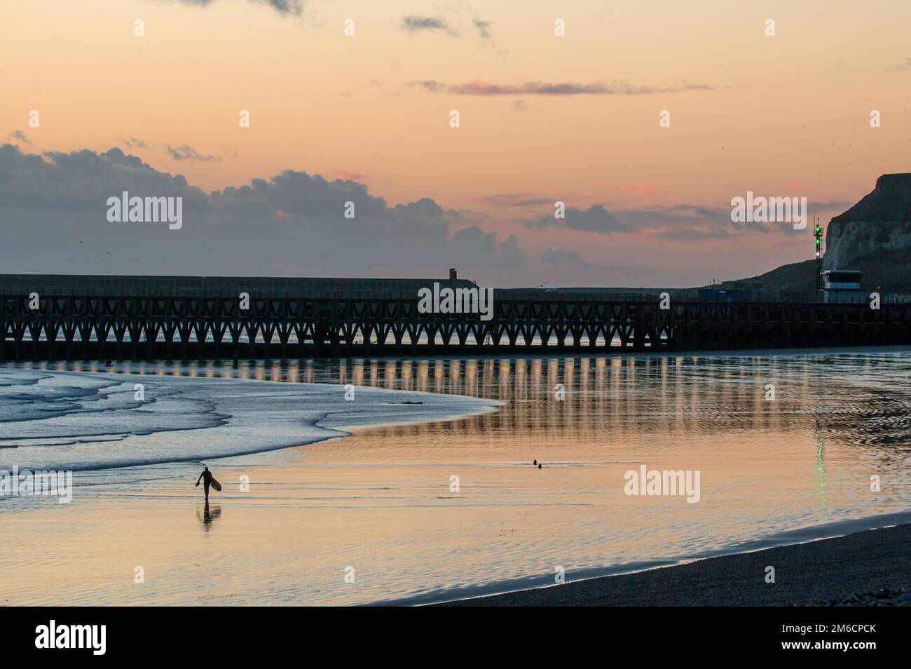 Persona non identificabile con tavola da surf a piedi in bassa marea a Seaford Beach al tramonto Foto Stock