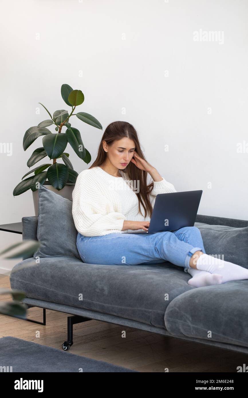 Un'attraente donna freelance è seduta sul divano e utilizza un computer portatile per lavorare online. Foto Stock