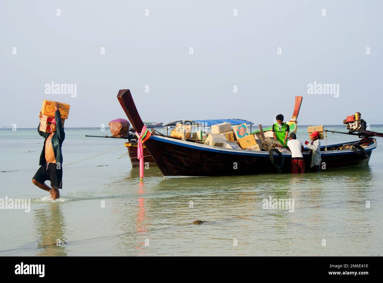 Persone che trasportano merci in scatole sulla costa dell'isola di Koh Lipe in Thailandia, barche di legno con colorate bande di tessuto arcobaleno, un soleggiato rilassante da Foto Stock