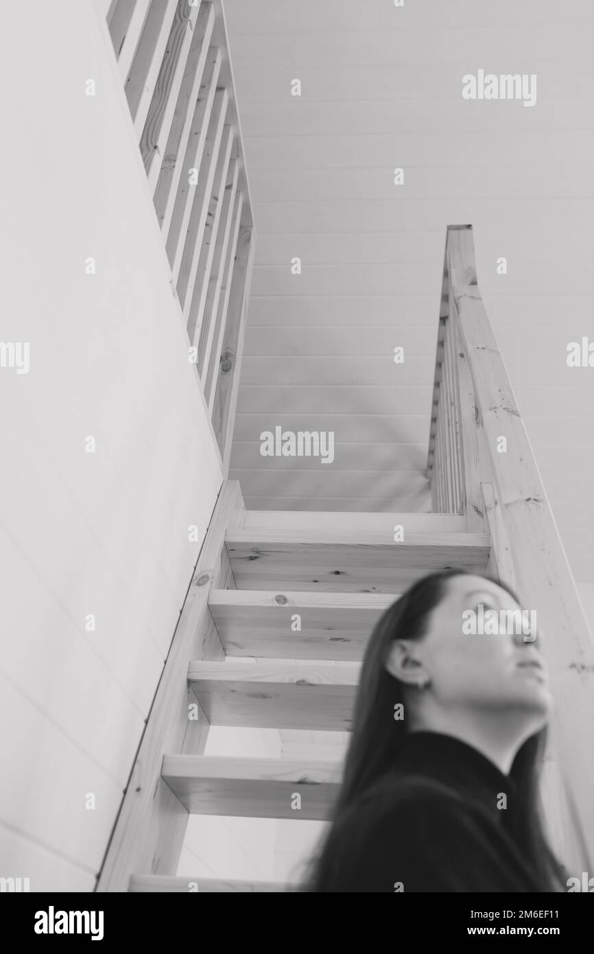 Giovane donna acconciatura semplice contro scale di legno. Depressione, solitudine e concetto di quarantena. Salute mentale, cura di sé, stare a casa. Nero e. Foto Stock