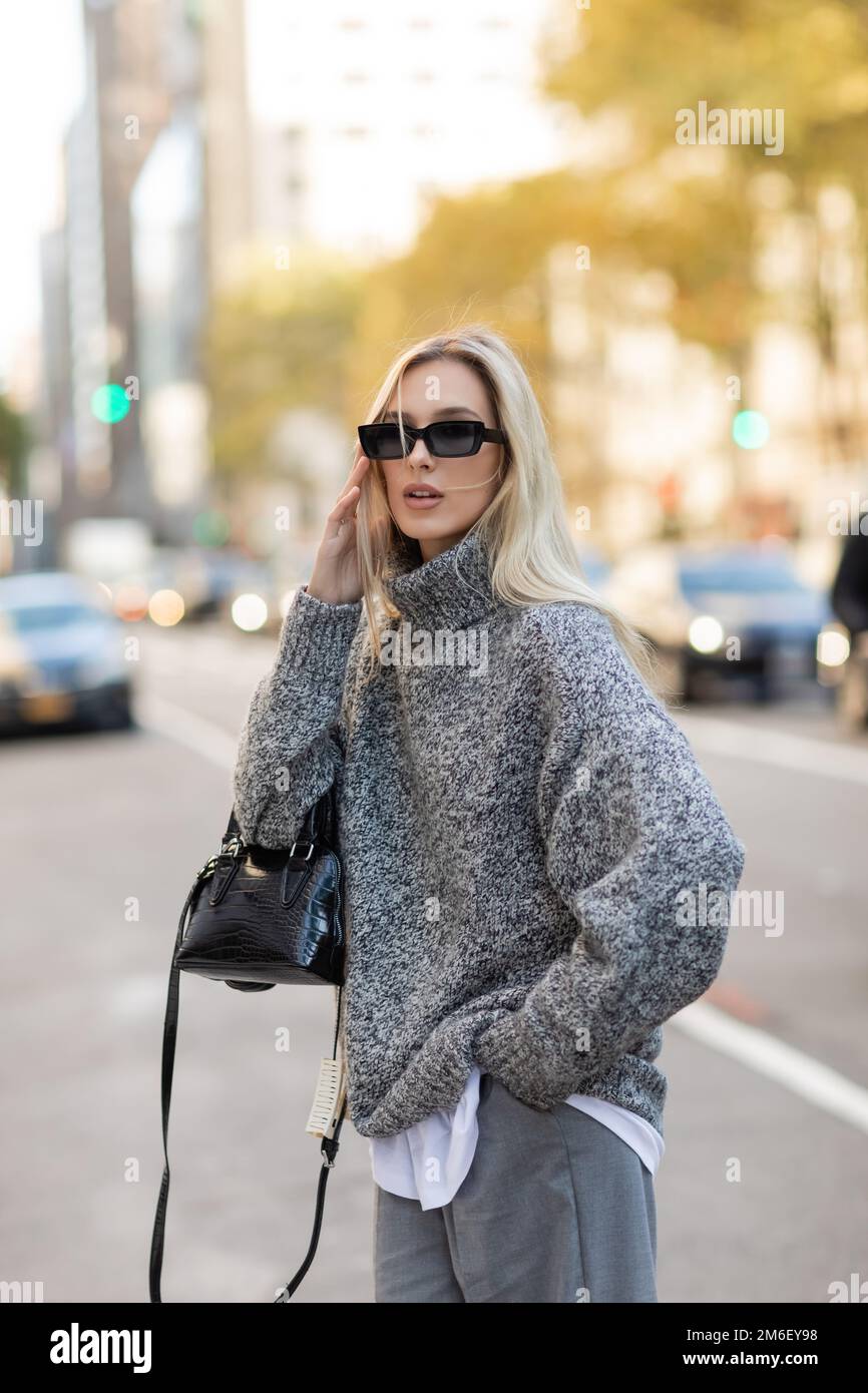 Donna bionda in maglione grigio che tiene la borsa e in piedi con la mano  in tasca sulla strada di New York City, immagine stock Foto stock - Alamy