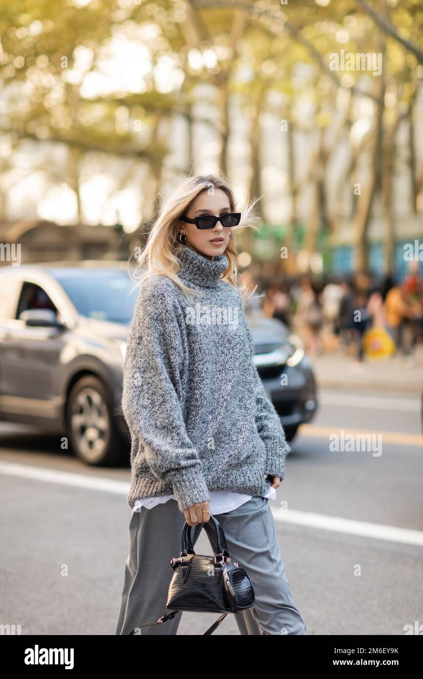 Donna alla moda in maglione grigio e occhiali da sole che tiene la borsa  mentre cammina per strada della città di New York, immagine stock Foto  stock - Alamy