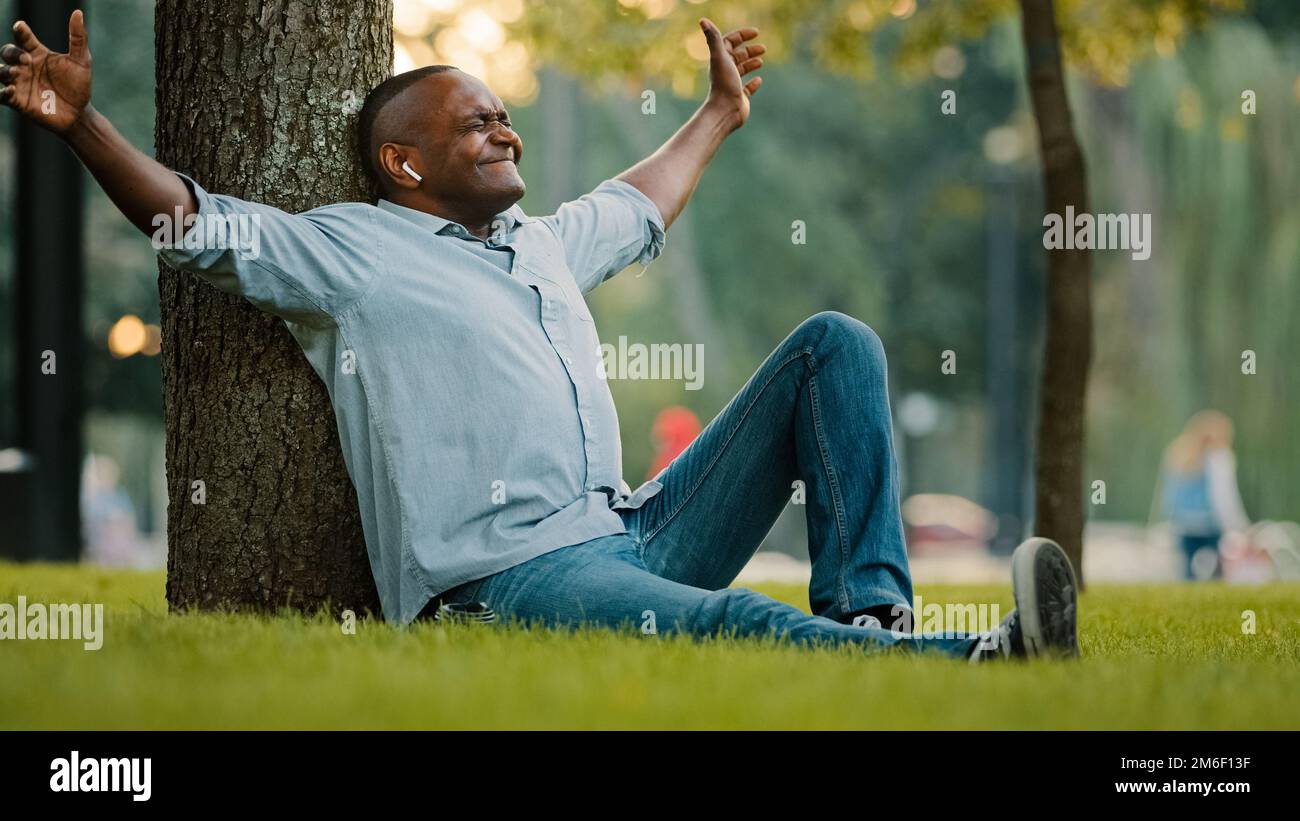Uomo d'affari afroamericano di mezza età in cuffie wireless seduto sull'erba nel parco cittadino uomo etnico ascoltare musica in auricolari allegri Foto Stock