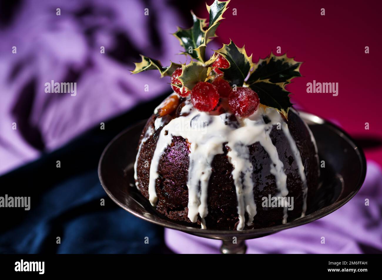 Tradizionale dessert natalizio al budino di frutta, decorato con ciliegie e foglie, con un classico sfondo rosso, blu e viola Foto Stock