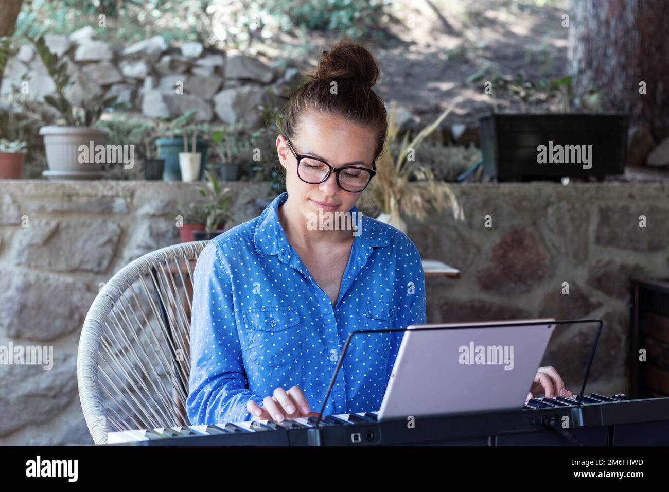 Giovane donna in occhiali che suona il pianoforte elettrico sulla terrazza utilizzando un tablet digitale per tutorial online. Foto Stock