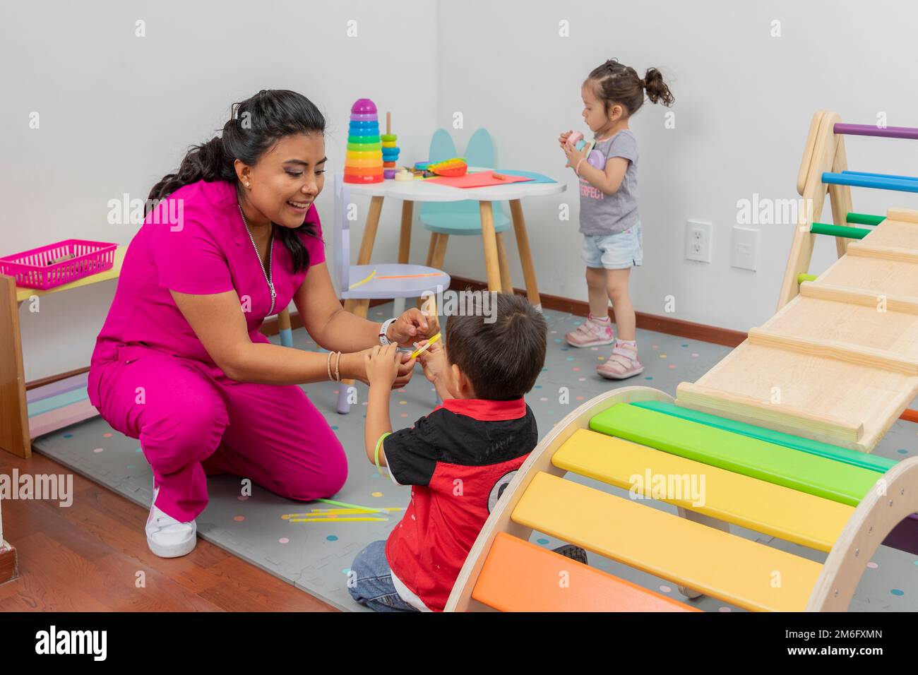 Pediatra latina che gioca con un bambino nel suo ufficio medico, mentre una ragazza gioca su un tavolo sullo sfondo. Foto Stock