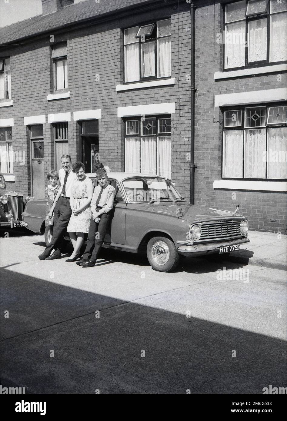 1960s, storico, una famiglia in piedi per una foto dalla loro Vauxhall Victor FB auto in una strada fuori la loro casa a schiera, Inghilterra, Regno Unito. Un'auto MG parcheggiata è dietro. Foto Stock