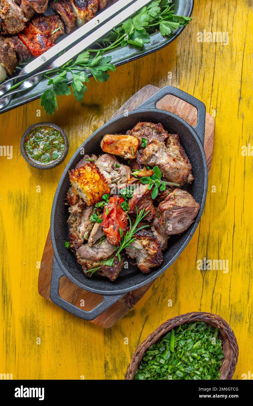 Grigliate di carne - maiale, pollo e verdure in padella di ghisa. Spiedini alla griglia su sfondo di legno giallo. Vista dall'alto. Foto Stock