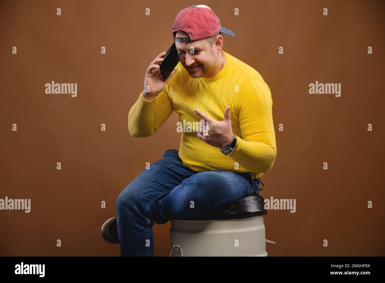 Un uomo parla in modo espressivo al telefono mentre si siede su un barile in studio. Chubby uomo che parla emotivamente sul suo smartphone Foto Stock