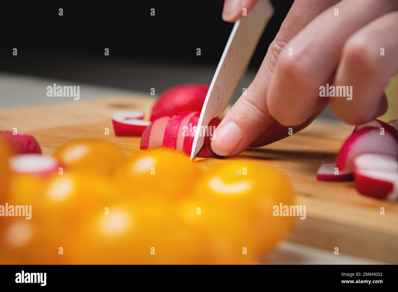 Primo piano delle mani femminili di rafano crudo fresco tagliato su un asse di legno sullo sfondo di pomodori ciliegini gialli. Tas vegetariano Foto Stock