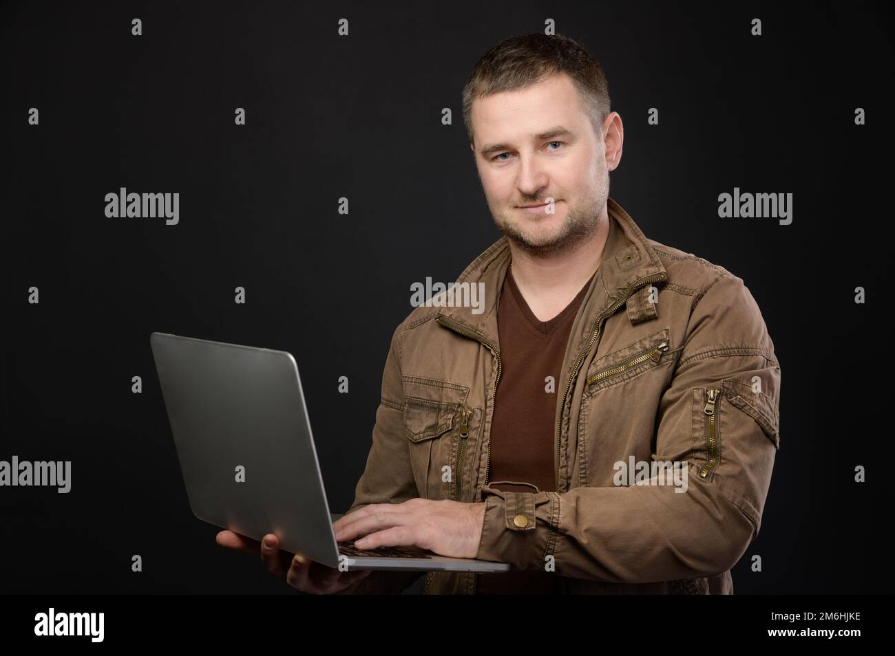 Ritratto di amichevole uomo casual caucasico positivo che utilizza un computer portatile grigio mentre si è in piedi in studio e guardando la fotocamera Foto Stock