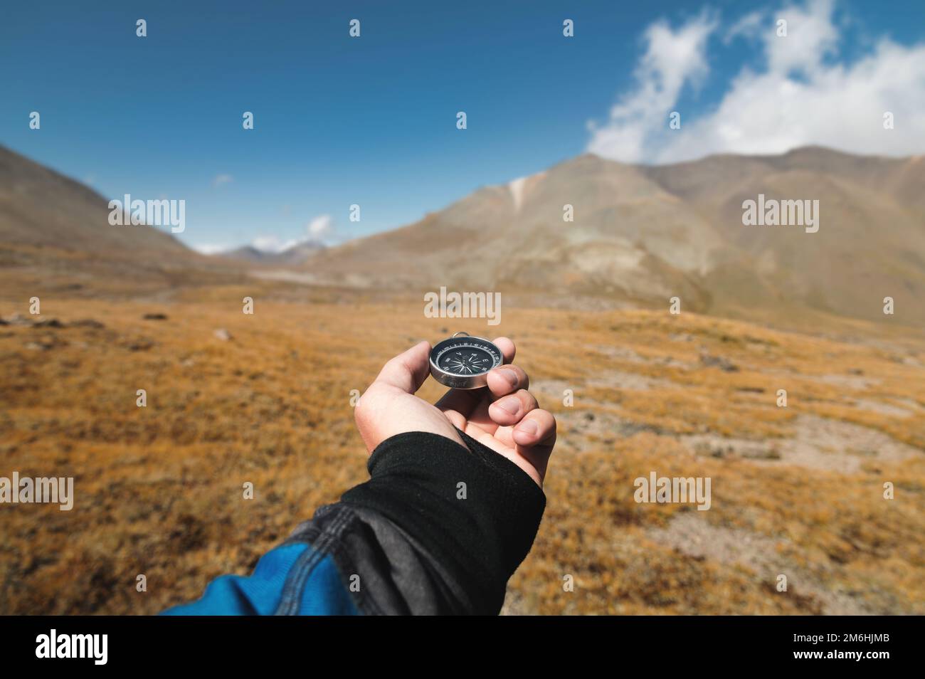 La mano di un viaggiatore maschio POV tiene una bussola magnetica sullo sfondo di un'area montuosa. Orientamento e ricerca Foto Stock
