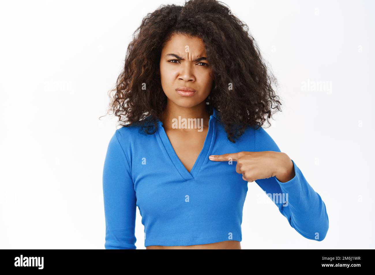 Ragazza nera arrabbiata, indicando se stessa e accigliandosi con il volto deluso, in piedi su sfondo bianco Foto Stock
