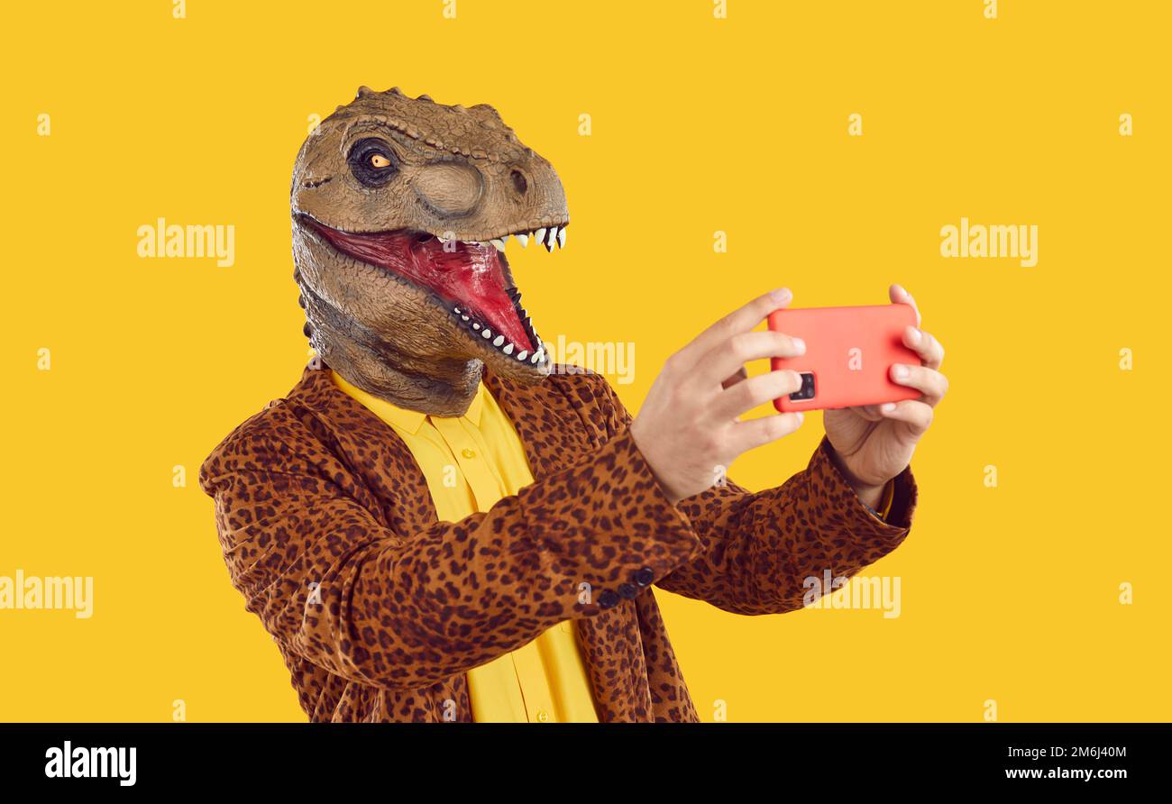 Eccentrico divertente uomo in gomma dinosauro maschera scattare foto selfie su smartphone su sfondo giallo. Foto Stock