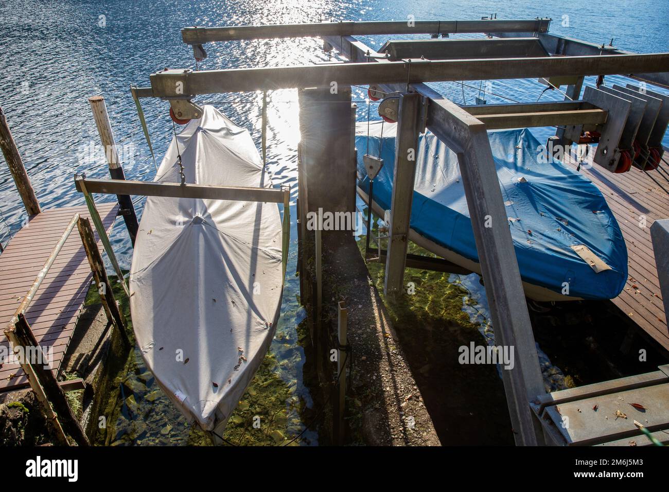 2 barche coperte e ora sollevate su un verricello. Vista dall'Olive Trail (un sobborgo di Lugano). I raggi del sole si riflettono nell'acqua del Lago di Lugano. Foto Stock