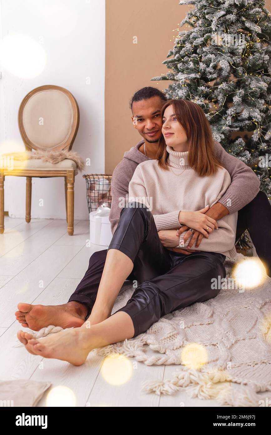 Un ragazzo di stile in una felpa marrone è seduto abbracciando la sua ragazza, vicino all'albero di Natale Foto Stock