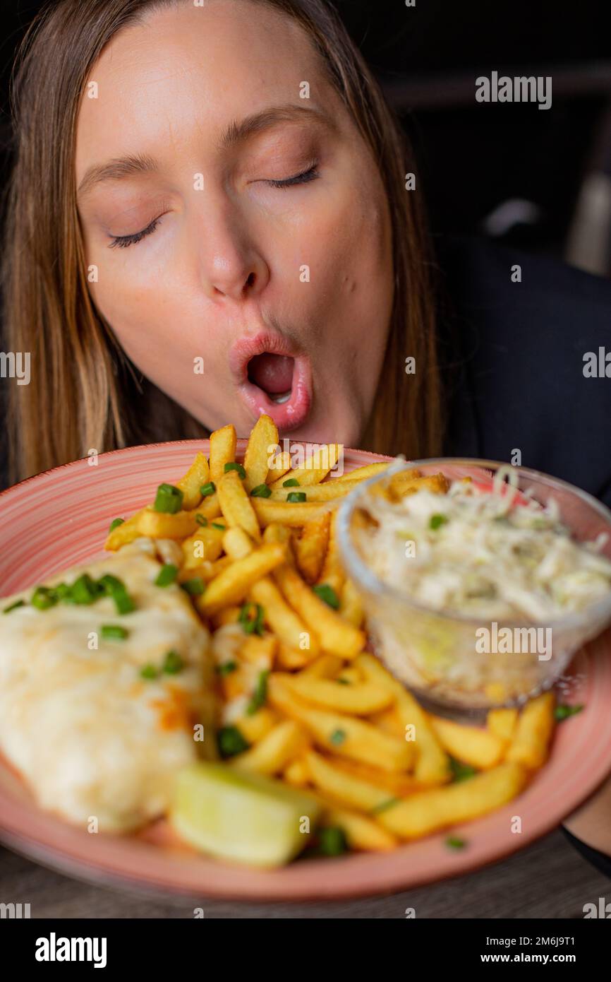 Giovane donna tenere piatto con patatine fritte, filetto di pollo e insalata di verdure e gustare il sapore con gli occhi chiusi e bocca aperta. Servizio di piatto principale e. Foto Stock