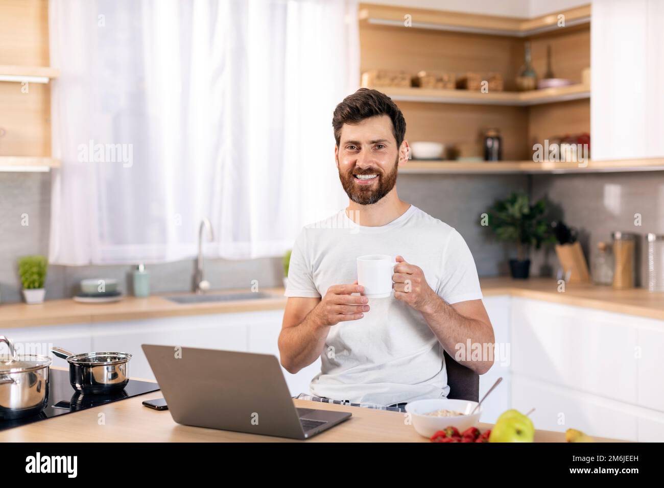 Sorridente maschio caucasico millenario con barba con tazza di caffè gode buona mattina, ha la colazione da solo con il computer Foto Stock