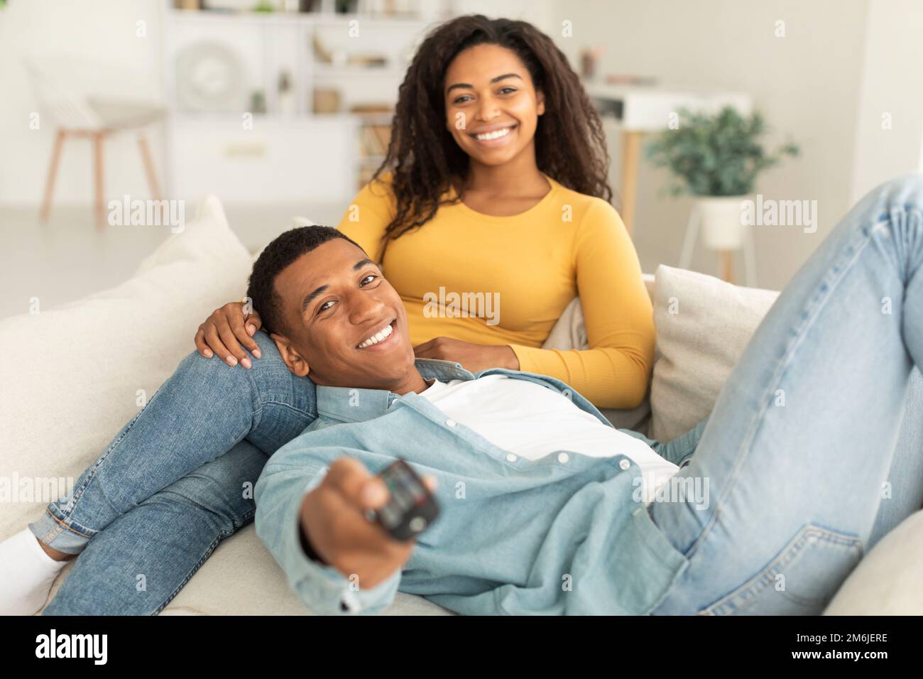 Allegro uomo afroamericano millenario con telecomando e donna godere del tempo libero, rilassarsi sul divano Foto Stock
