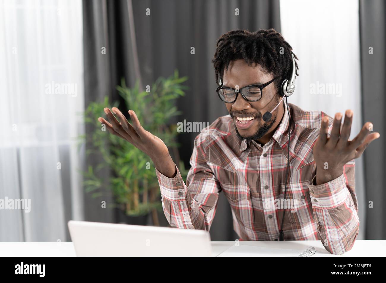 Freelance afro-americano gesturing guardando il laptop con le cuffie che hanno una videochiamata di gruppo che lavora lontano a casa. Tu Foto Stock