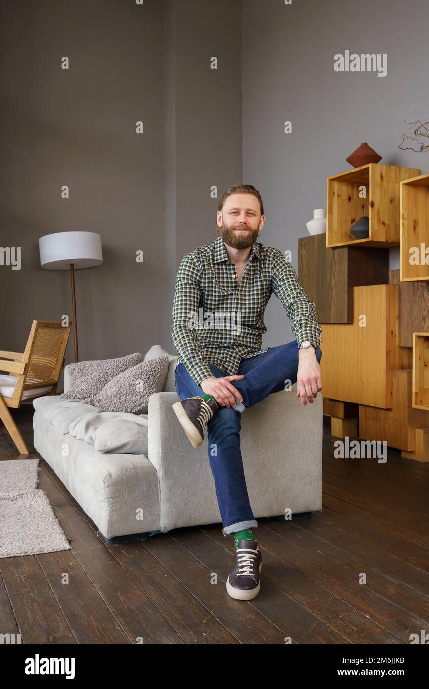 Un bell'uomo di mezza età si siede sul divano del soggiorno, ha una barba e capelli lunghi, è vestito di jeans e. Foto Stock Un bell'uomo di mezza età si siede sul divano del soggiorno, ha una barba e capelli lunghi, è vestito di jeans e. Foto Stock