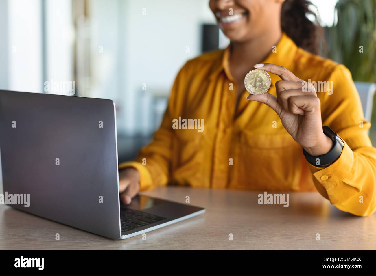 Commercio di criptovaluta. Donna d'affari nera irriconoscibile seduta alla scrivania con il laptop e che mostra il bitcoin dorato Foto Stock
