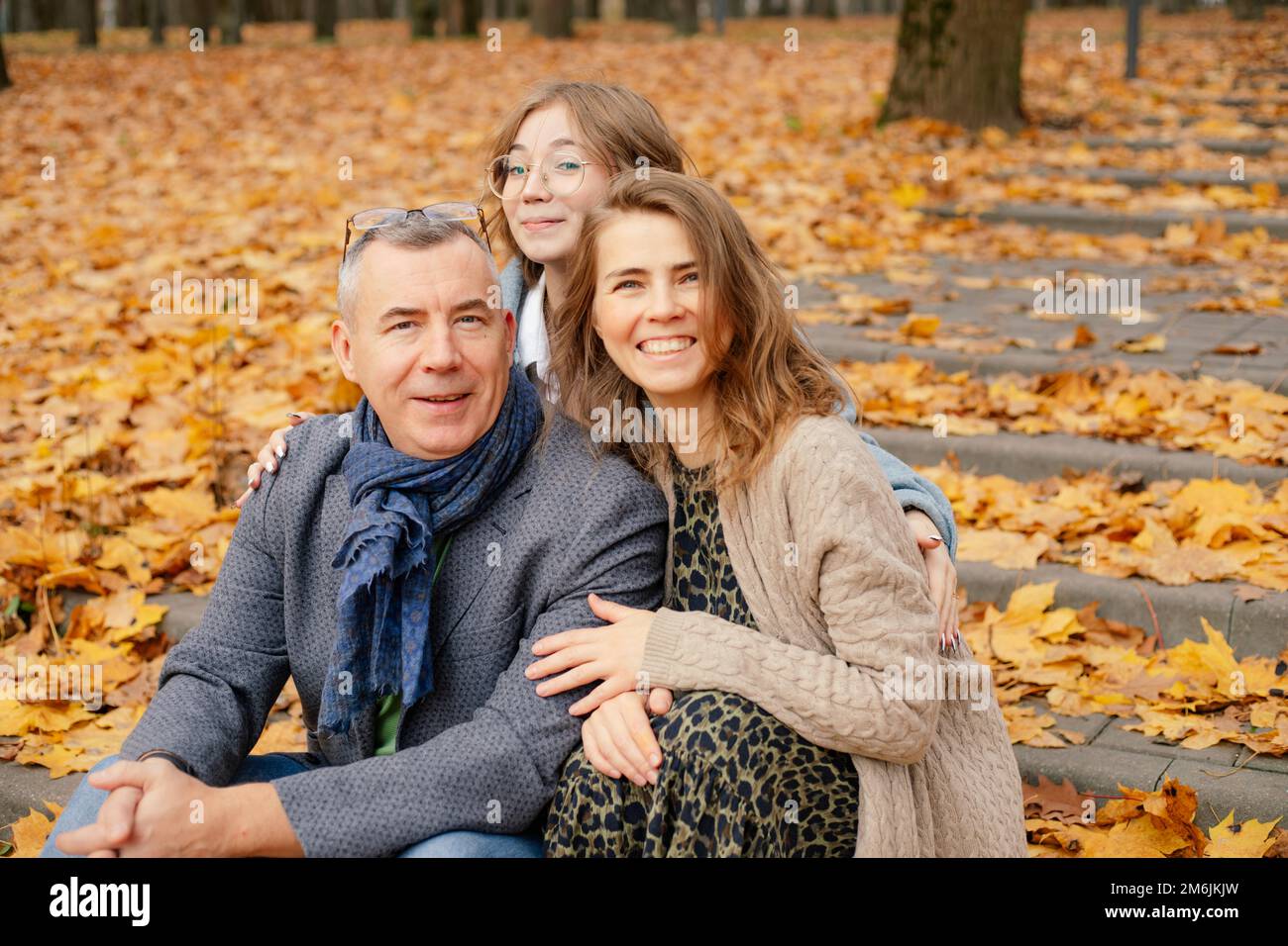 Ritratto di famiglia abbracciante, seduto su scale di cemento coperto di foglie gialle cadute tra gli alberi nel parco foresta. Foto Stock