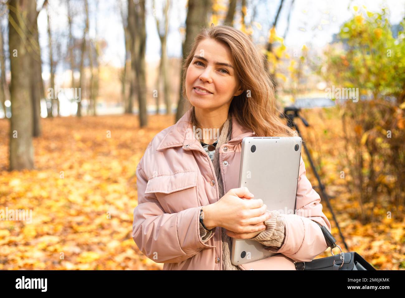 Ritratto di donna di mezza età in piedi vicino agli alberi su foglie di acero giallo caduto nella foresta del parco in autunno tenendo un computer portatile. Foto Stock