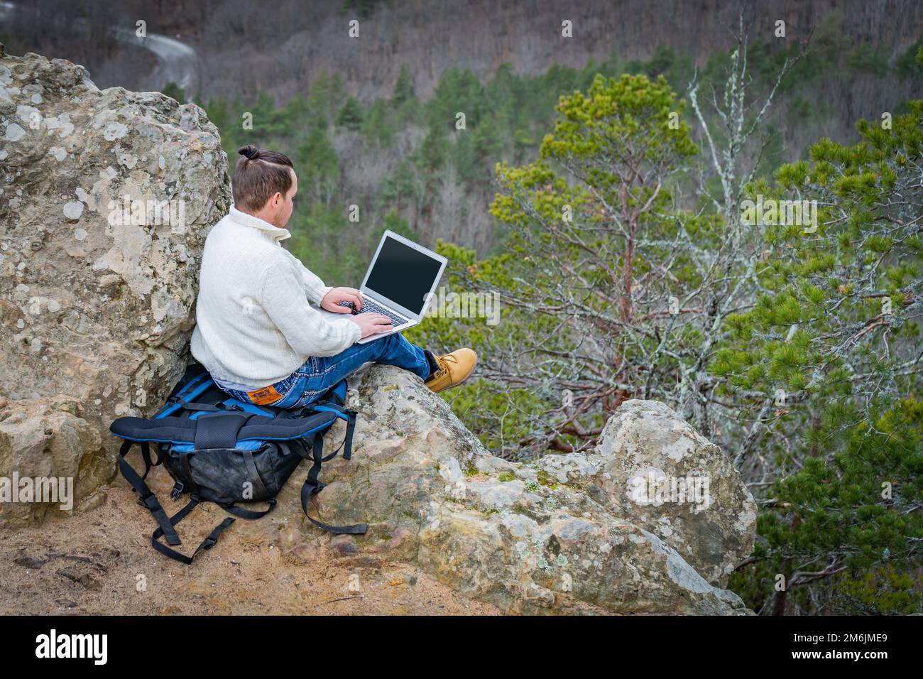 Un uomo di viaggio che lavora con un computer portatile seduto sulla montagna rocciosa sullo sfondo di una splendida scogliera panoramica Foto Stock