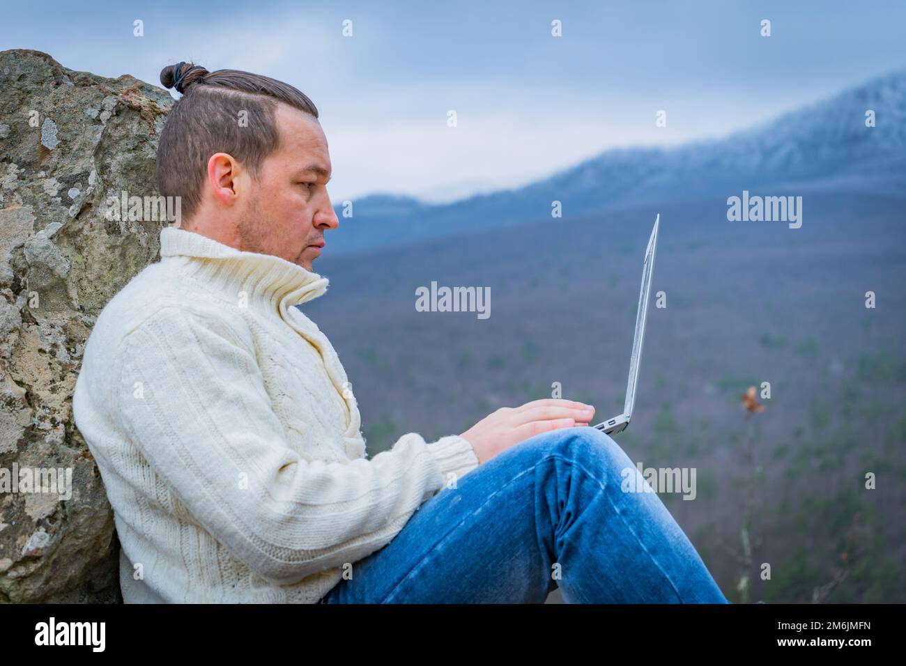 Uomo hipster che lavora con un computer portatile seduto sulla montagna rocciosa su uno splendido sfondo scenica della scogliera Foto Stock