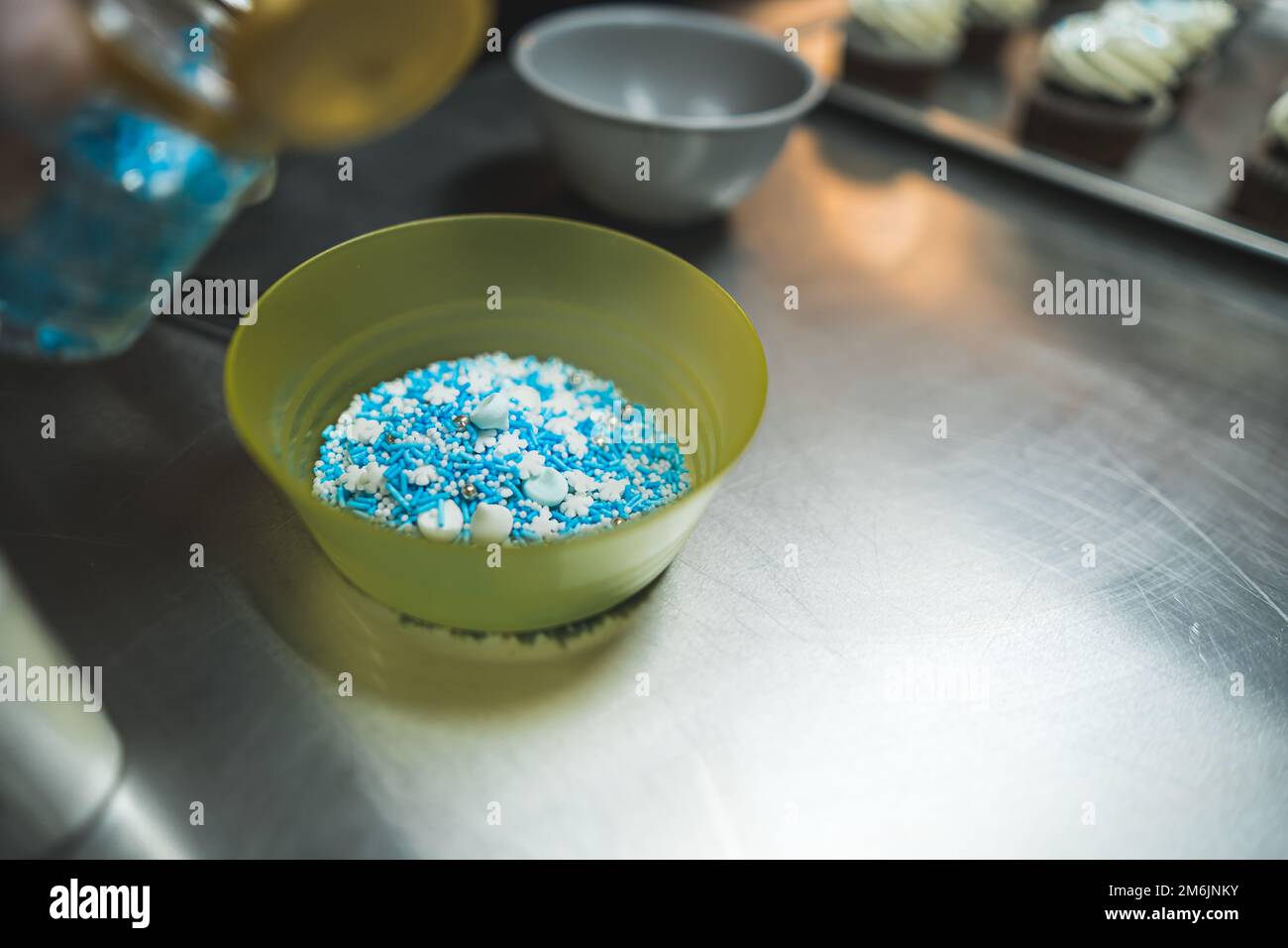 Spruzzare blu in una ciotola gialla. Vaso aperto con spruzzi blu sfocati in primo piano. Cupcake decorati con crema bianca posta su una teglia da forno sfocata sullo sfondo. Foto di alta qualità Foto Stock