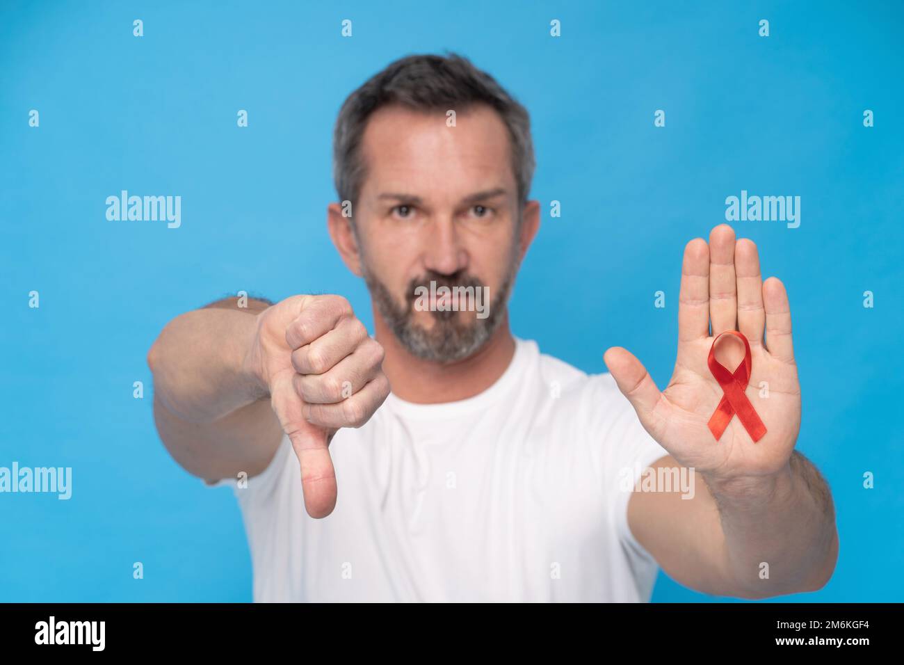 Uomo maturo che gestura il pollice giù con il nastro rosso fiocco AIDS simbolo di consapevolezza su una palma che indossa una t-shirt bianca isolata su una b blu Foto Stock