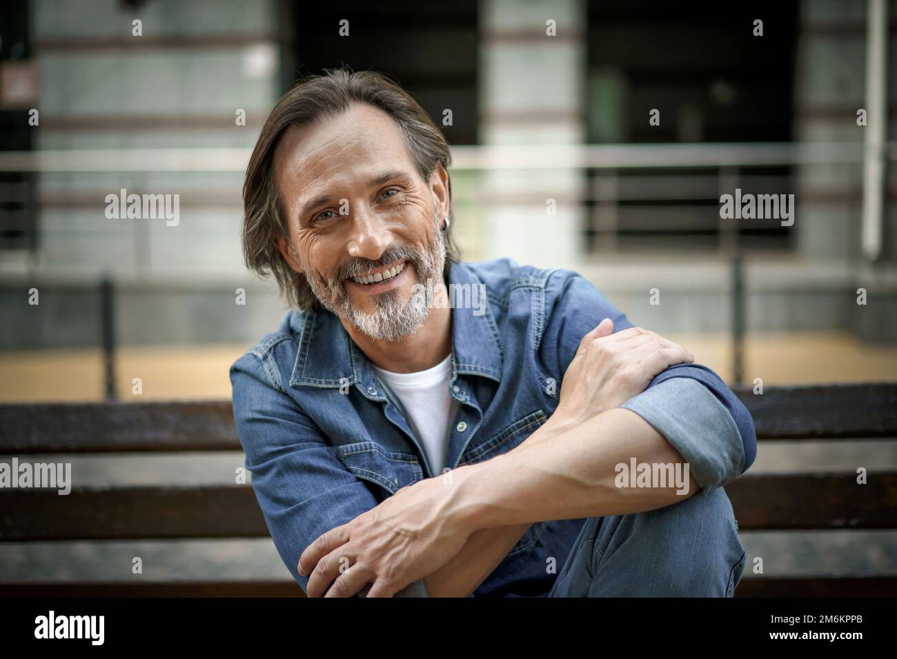 Felice sorridente bella barba grigia uomo di mezza età si siede alla panca con le mani ripiegate appoggiate sul ginocchio sollevato all'aperto in città cit Foto Stock