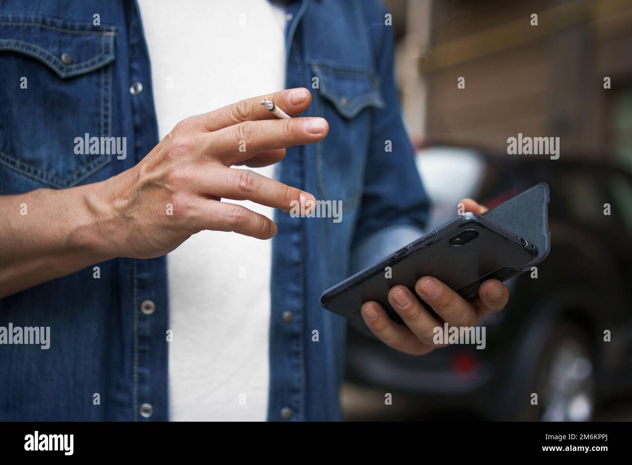 Fumo sigaret uomo di mezza età che controlla messaggi di testo o social media utilizzando il suo smartphone indossando jeans camicia e t-shir bianco Foto Stock