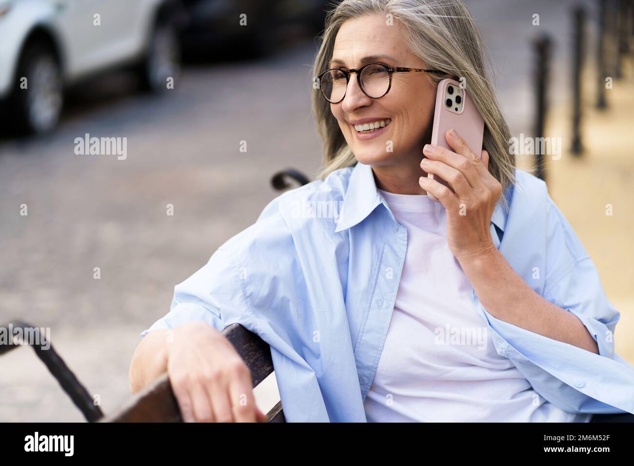 In estate, donna matura con i capelli grigi che parlano sul telefono che tiene lo smartphone seduto sulla panca per le strade di Foto Stock