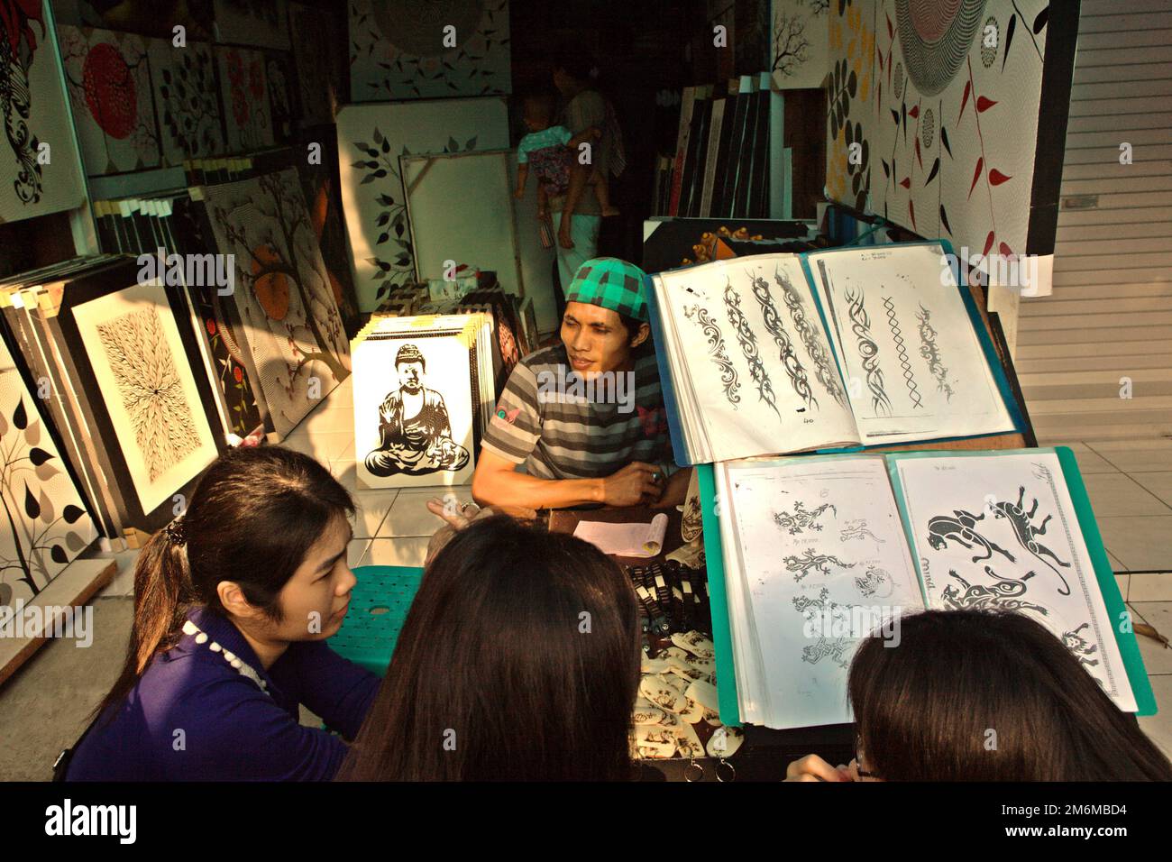 Un acquirente aspetta come un gruppo di turisti donne stanno discutendo le opzioni mentre accovacciandosi davanti ad un grande libro di esposizione di disegno del tatuaggio ad un negozio di arte del streetside a Tanah Lot, Tabanan, Bali, Indonesia. Foto Stock