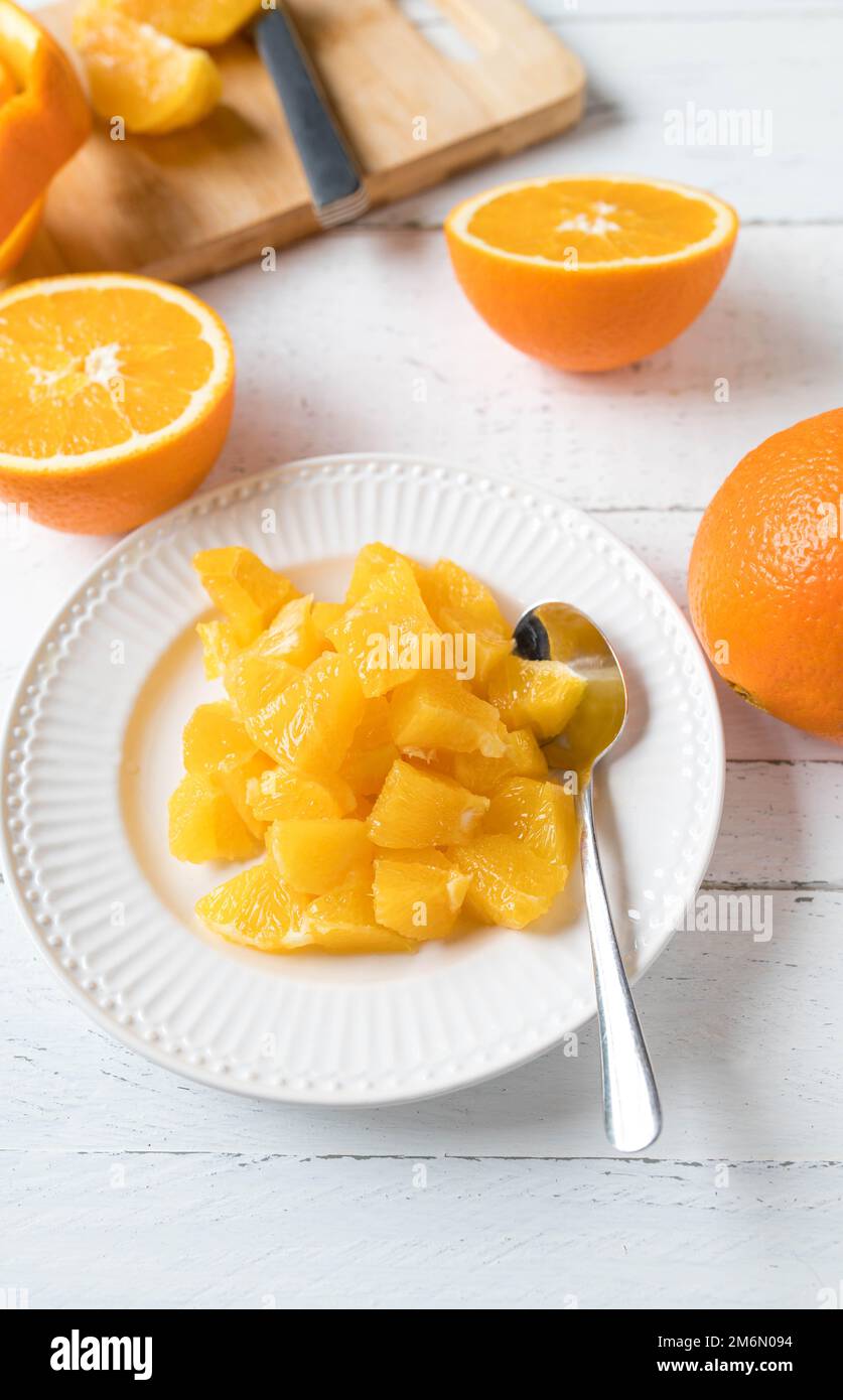 Arance tagliate a dadini o tritate su un piatto per uno spuntino sano o dessert su sfondo di legno bianco con tagliere, buccia e coltello Foto Stock