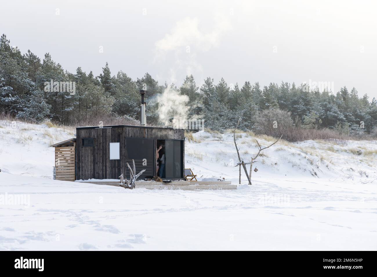 Sauna all'aperto in legno con fumo che esce dal camino in una bella giornata invernale fredda e innevata al Mar Baltico. Benessere e stile di vita sano. Foto Stock