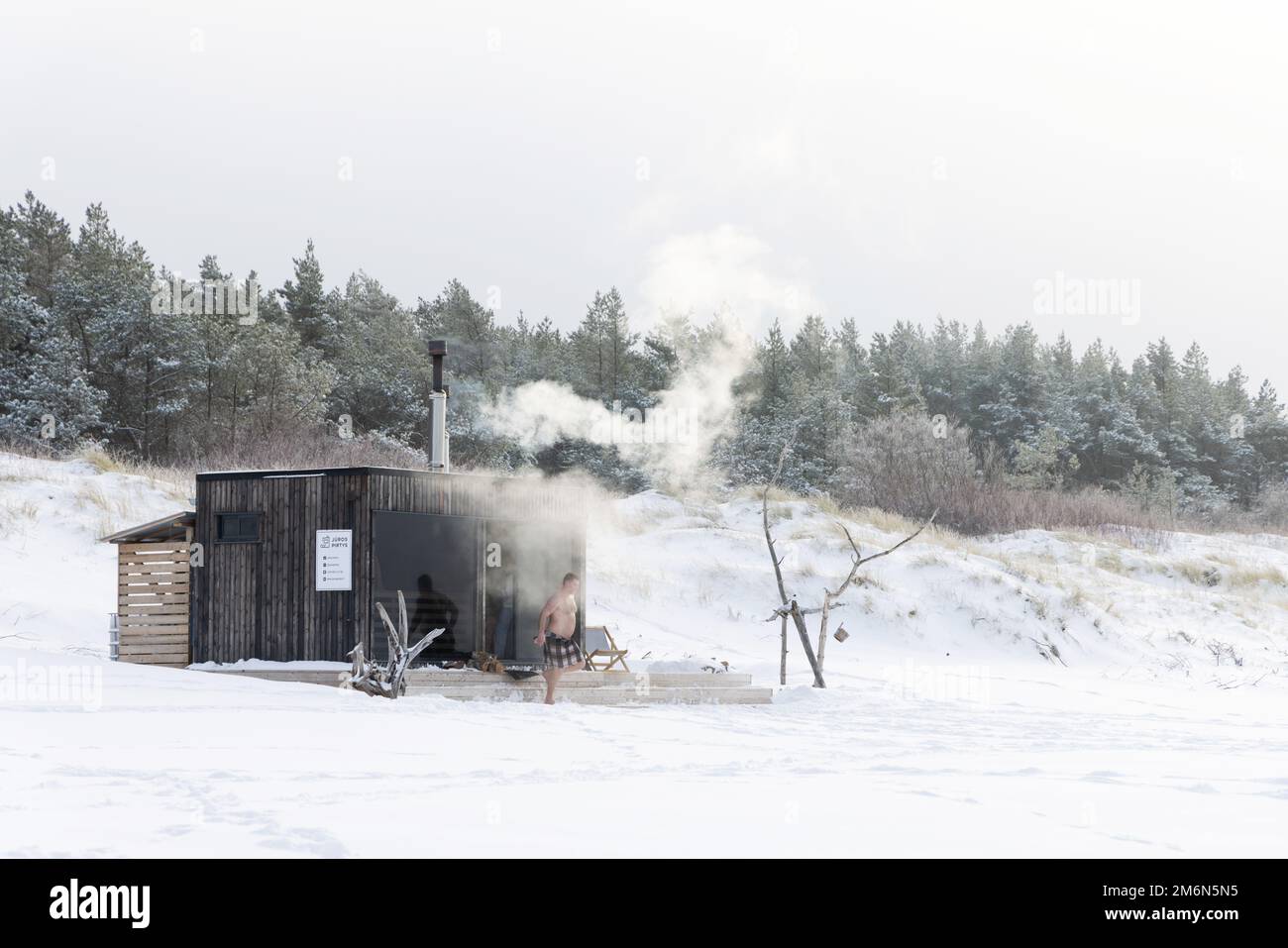 Sauna all'aperto in legno con fumo che esce dal camino in una bella giornata invernale fredda e innevata al Mar Baltico. Benessere e stile di vita sano. Foto Stock