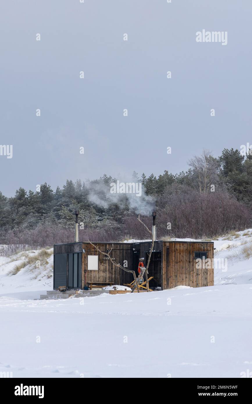 Sauna all'aperto in legno con fumo che esce dal camino in una bella giornata invernale fredda e innevata al Mar Baltico. Benessere e stile di vita sano. Foto Stock