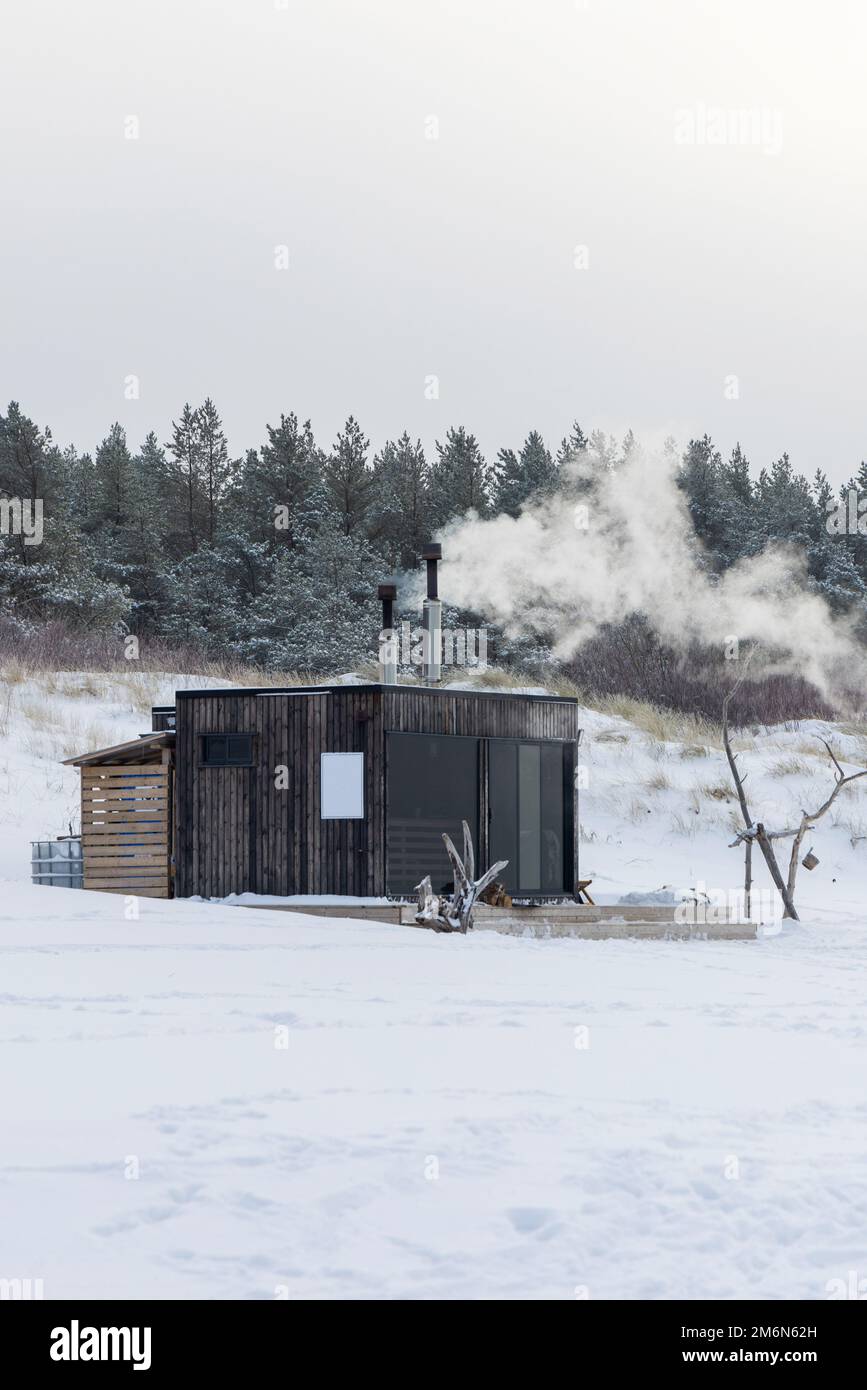 Sauna all'aperto in legno con fumo che esce dal camino in una bella giornata invernale fredda e innevata al Mar Baltico. Benessere e stile di vita sano. Foto Stock