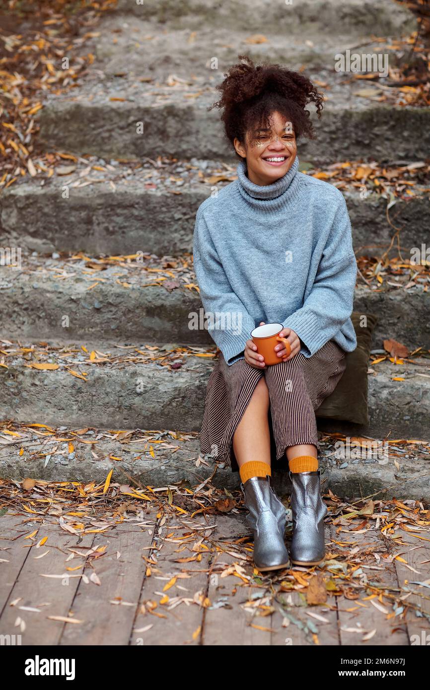 Bella giovane donna felice di etnia africana in abiti caldi seduti fuori il giorno d'autunno Foto Stock