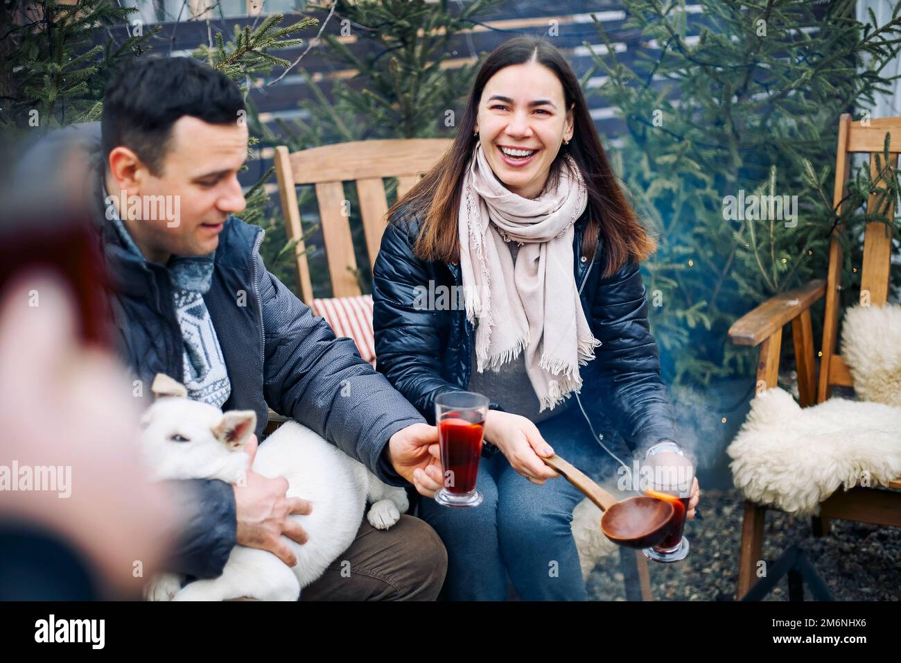 Festa invernale falò. Giovane coppia allegra con cane che beve VIN brulé caldo in compagnia di amici Foto Stock