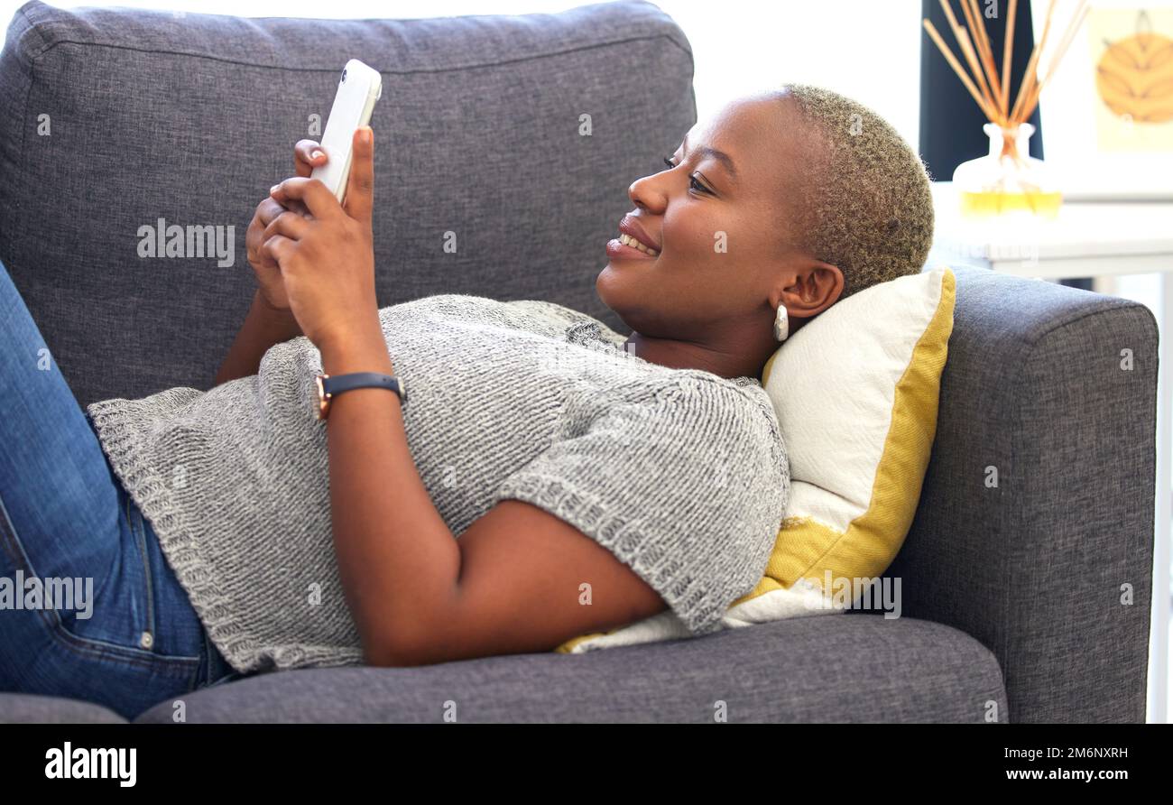 Social media, relax e donna nera con un telefono per la comunicazione, chat mobile e app sul divano. Connessione, internet e tranquilla ragazza africana con un Foto Stock