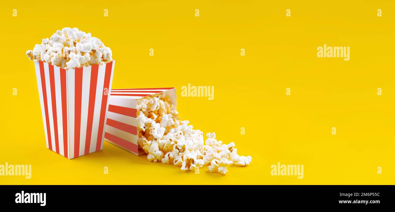 Due secchi in cartone a strisce bianche rosse con gustosi popcorn al formaggio, isolati su sfondo giallo. Scatola con dispersione di grani di popcorn. Film, cinema a. Foto Stock