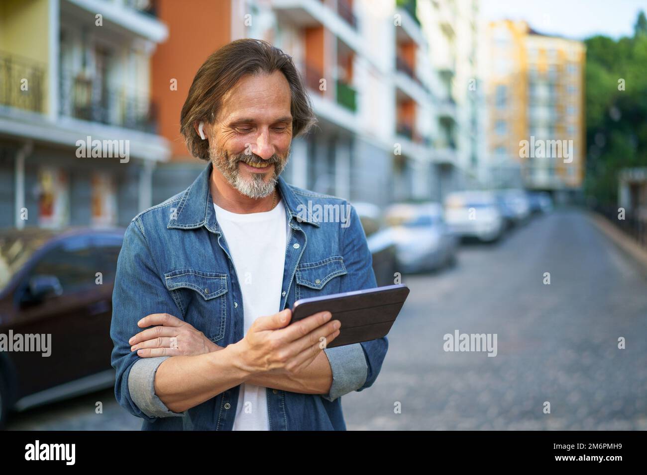 Un bell'uomo maturo in viaggio che guarda il tablet digitale in mano che parla all'aperto mentre chiama per le strade urbane. Uomo maturo Foto Stock