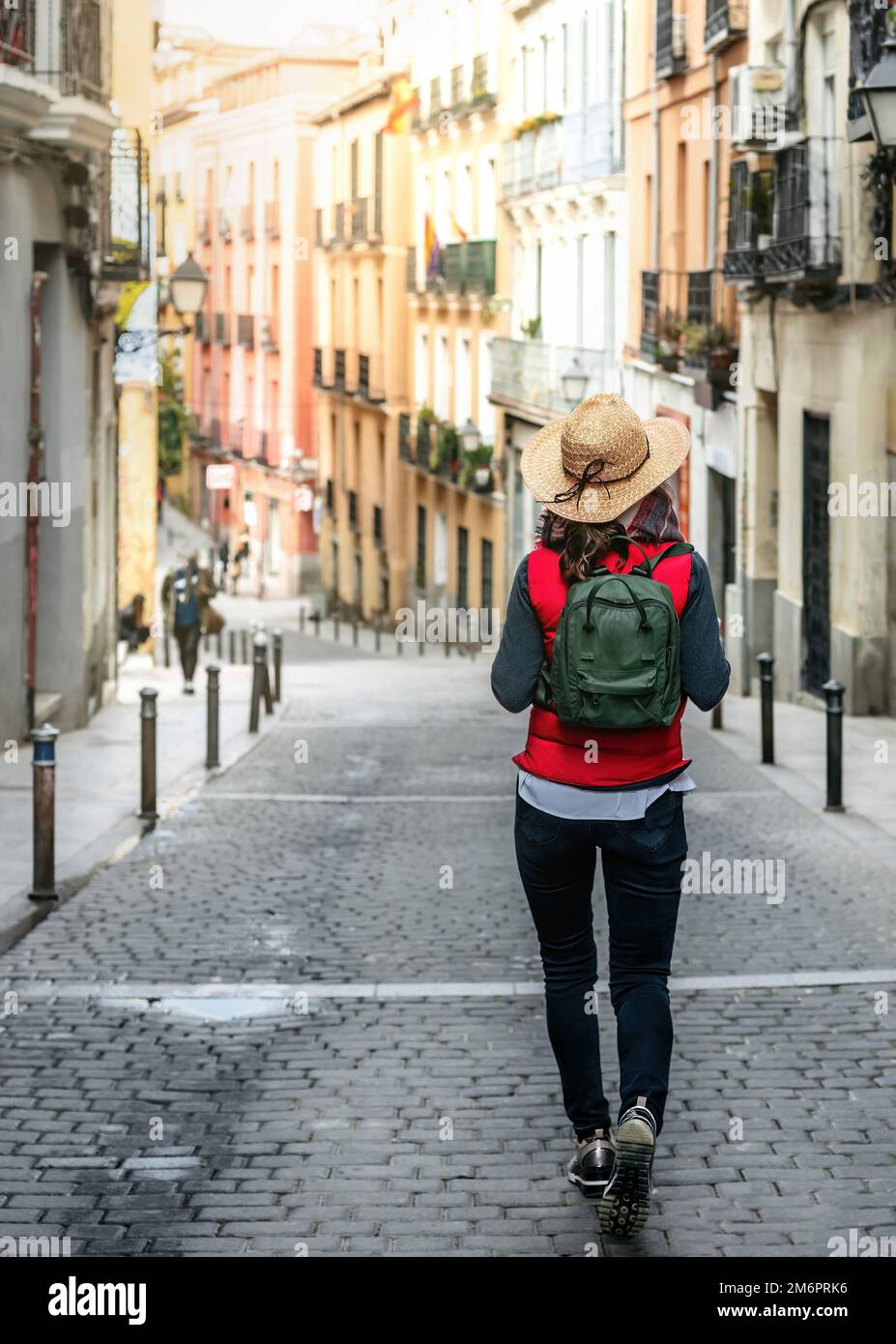 Una turista femminile con la schiena girata, indossando un cappello e uno zaino, cammina per una strada nel centro storico di Madrid. Turismo escursionistico in Spagna. Foto Stock