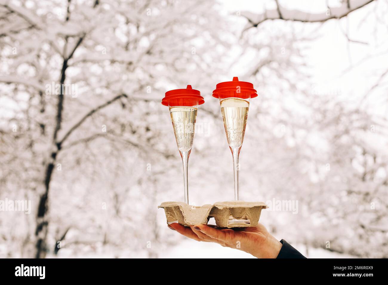 Donna anziana felice in abiti caldi che tiene due bicchieri di champagne mentre si trova in piedi nel cortile innevato Foto Stock